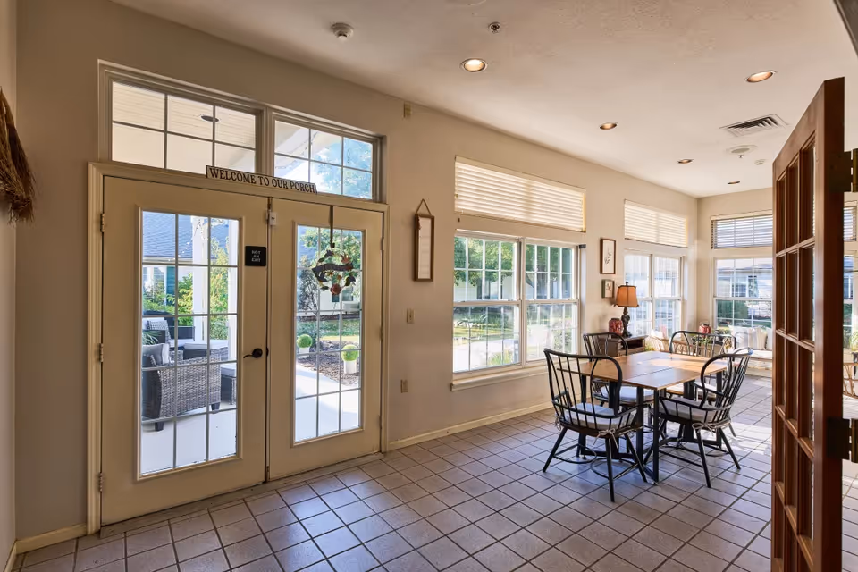 A bright interior room with tiled floor featuring a wooden table and four chairs with cushions. Large windows with blinds let in natural light. Double glass doors with a sign above reading 'WELCOME TO OUR PORCH' open to an outdoor seating area with wicker furniture and greenery.