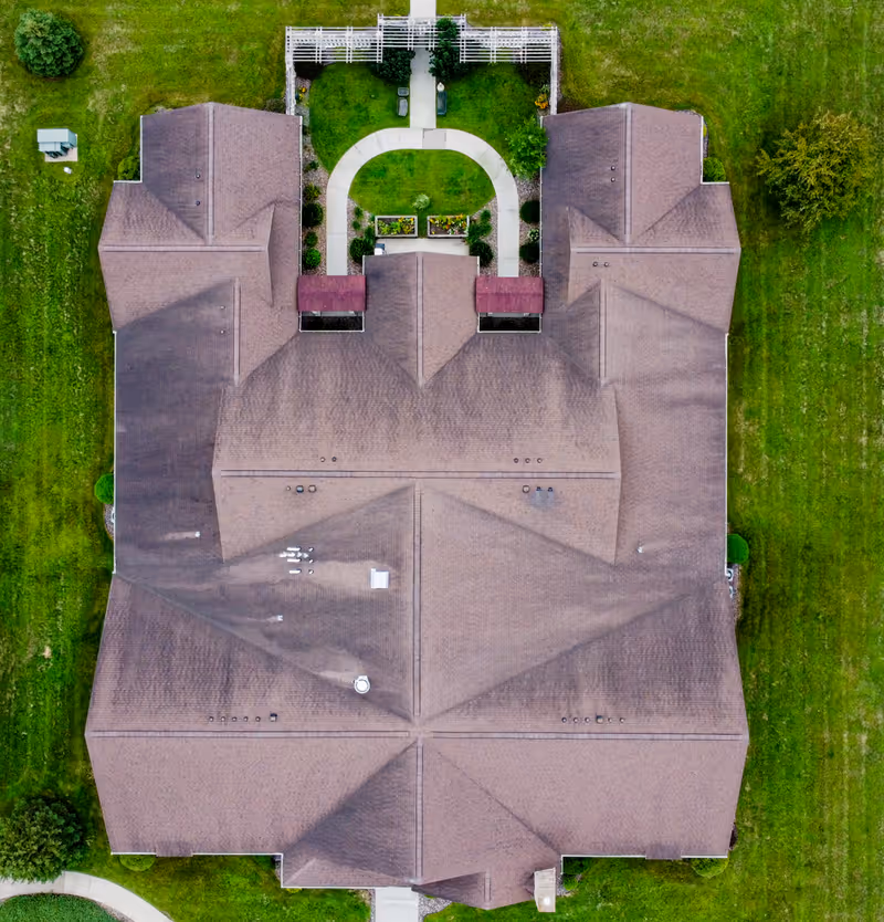 Aerial view of a large building with a brown roof surrounded by green grass and a small garden area with a white fence and pathways at the top center of the image.