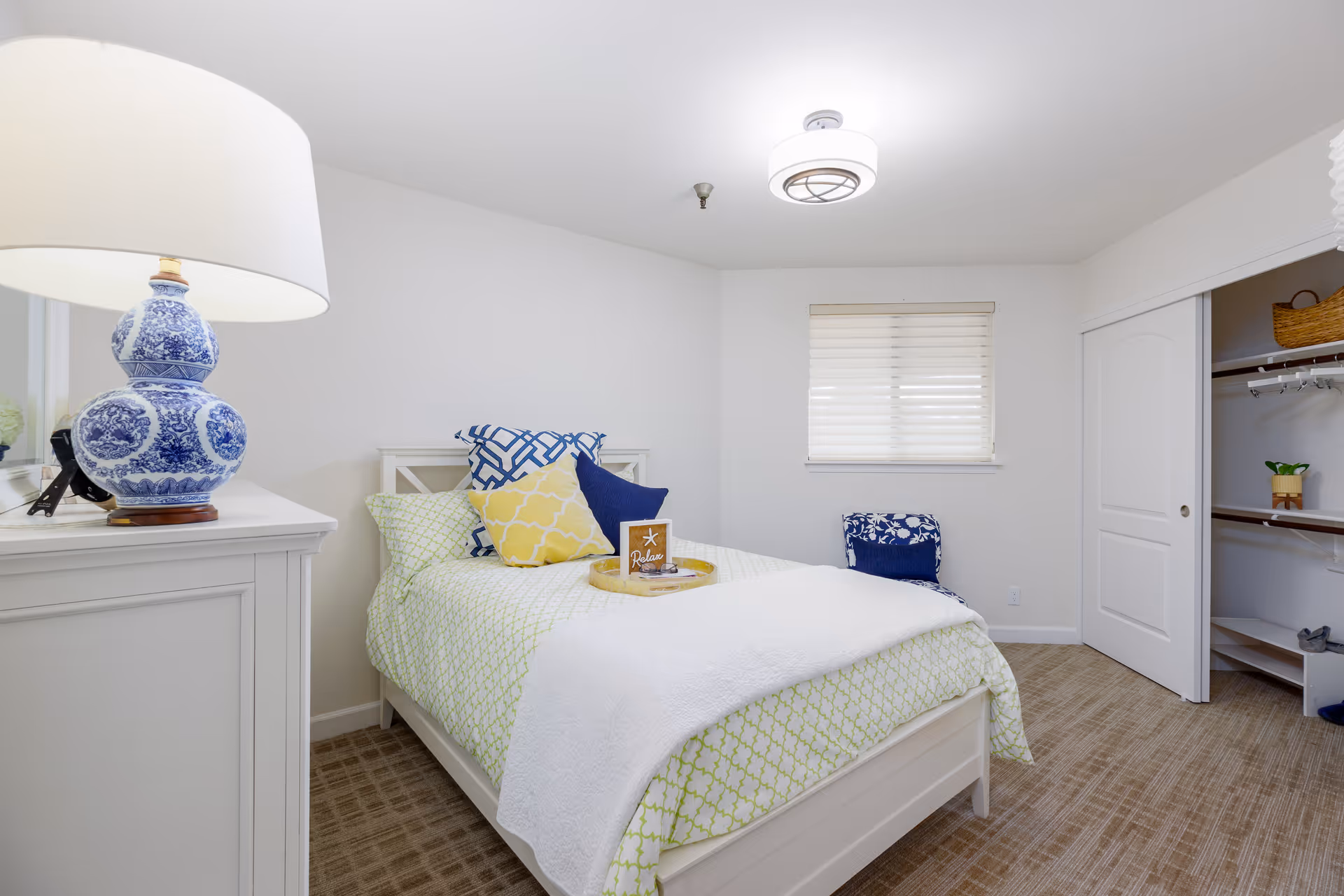 A bright and clean bedroom with a white bed frame and green patterned bedding. The bed is decorated with multiple pillows in blue, yellow, and white colors. A white dresser with a blue and white decorative lamp is on the left side. There is a window with blinds on the far wall and an open closet with sliding doors on the right. The room has beige carpet flooring and white walls.