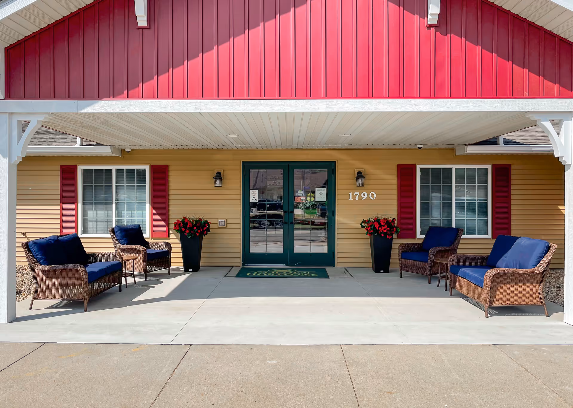 Entrance of a senior living facility with a covered porch area featuring two wicker sofas and two wicker chairs with blue cushions, two tall black planters with red flowers, beige siding with red shutters on windows, green double doors, and the number 1790 displayed on the wall.