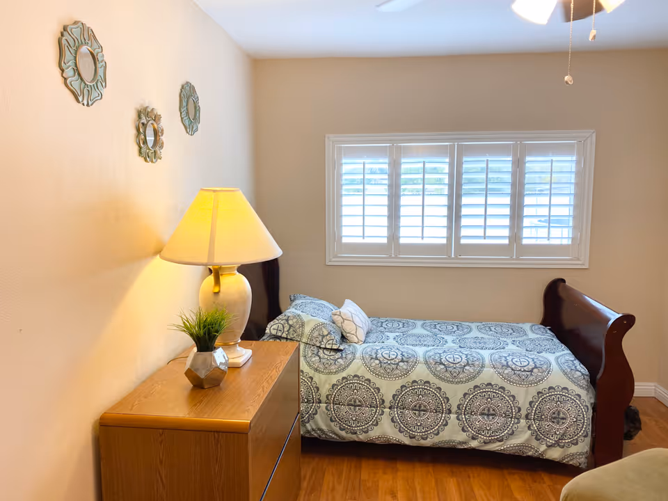 A small bedroom with a single bed covered in a patterned blue and white bedspread. Next to the bed is a wooden nightstand with a table lamp and a small potted plant. Three decorative mirrors are hung on the beige wall above the nightstand. A window with white shutters is on the wall behind the bed, allowing natural light into the room. The floor is wooden.