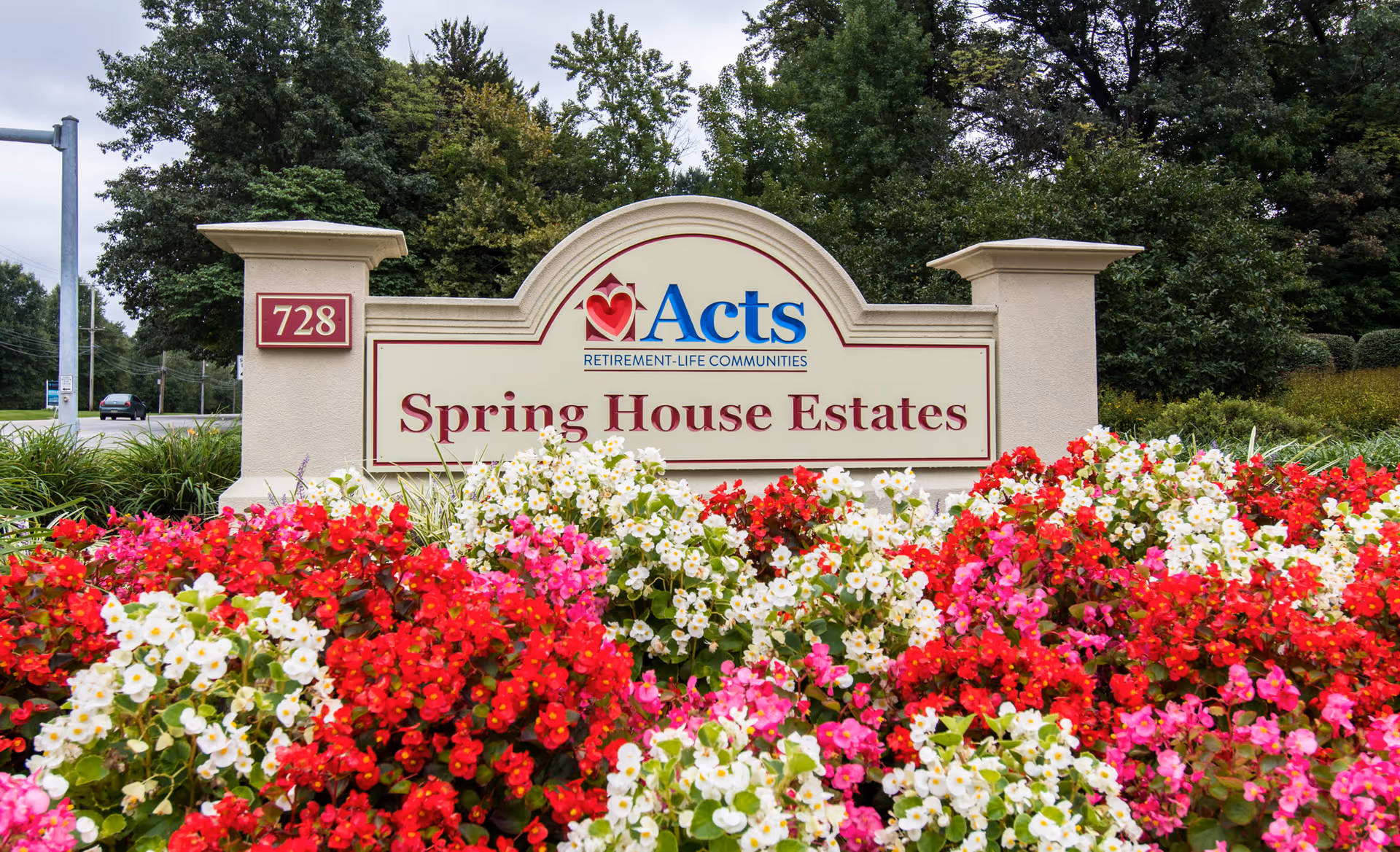 Entrance sign for Spring House Estates, a retirement-life community by Acts, surrounded by vibrant red, pink, and white flowers with trees and a road in the background.