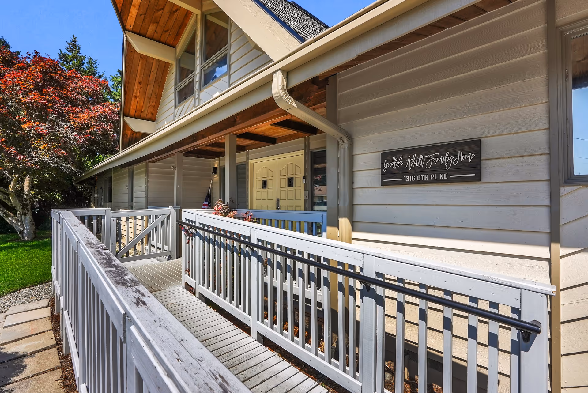 Exterior view of Goodlife Adult Family Home showing a wooden ramp with railings leading to double front doors under a covered porch. The building has beige siding and a sign with the facility name and address. There is a tree with red leaves and green grass on the left side.