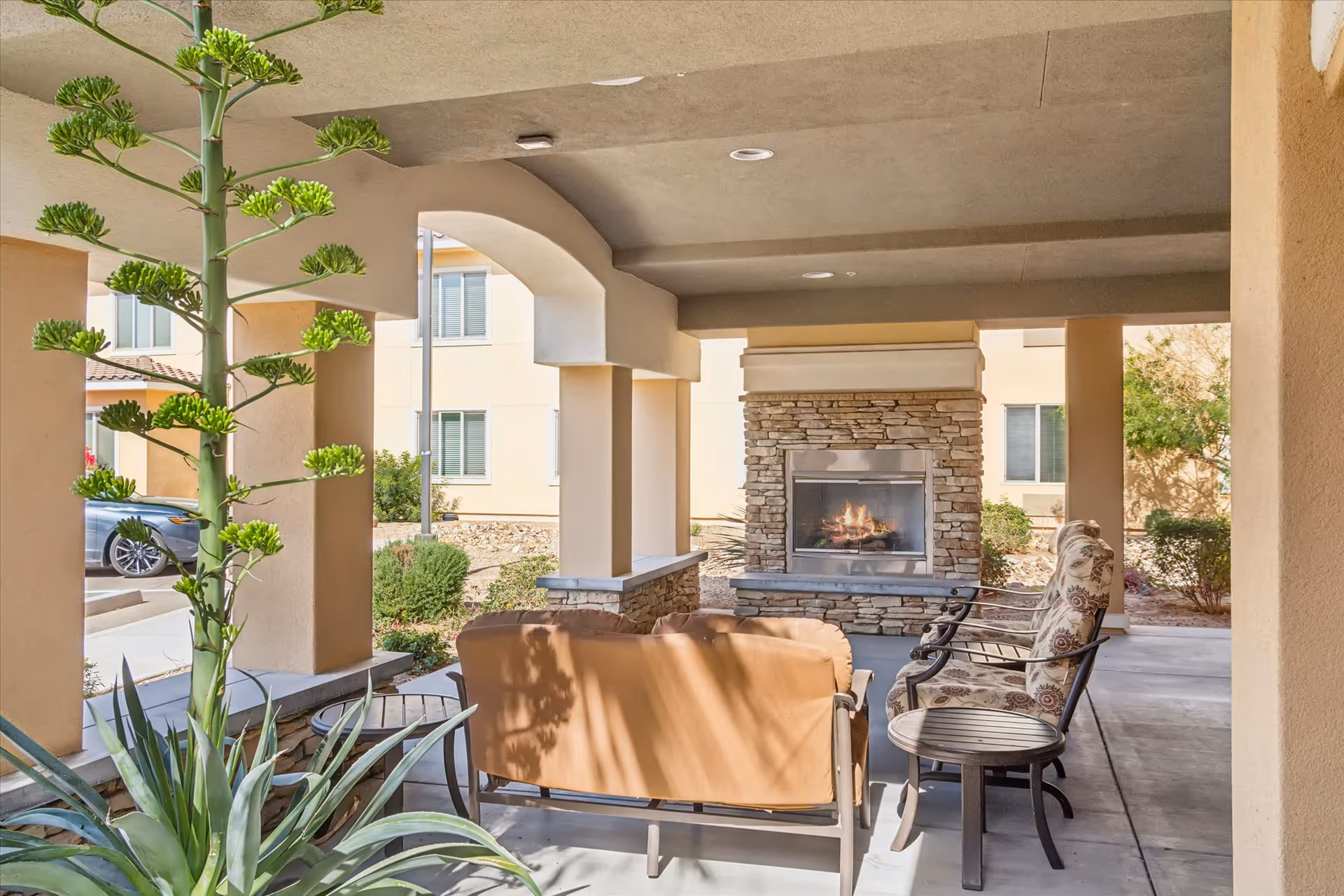 Covered outdoor seating area with cushioned chairs and a sofa arranged around a stone fireplace. The area is supported by beige columns and overlooks a garden with plants and shrubs. A car is partially visible in the background near the building.