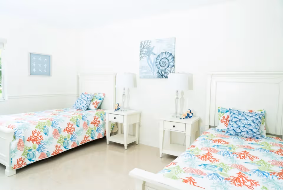 A bright bedroom with two white twin beds featuring colorful sea-themed bedding with coral and seashell patterns. Each bed has a matching pillow and a white nightstand with a lamp and small decorative items. The walls are white with two framed sea-themed artworks hanging above the beds.