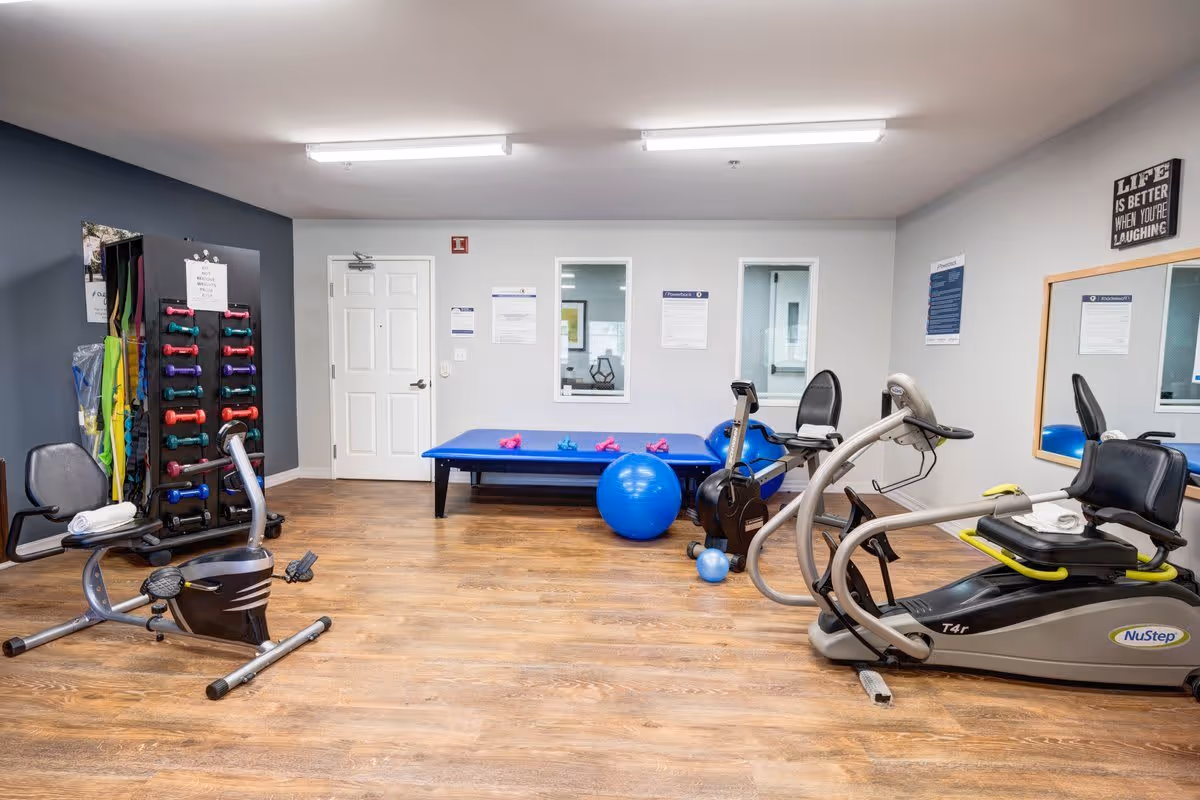 A fitness room in Creston Village with exercise equipment including a recumbent bike, a NuStep T4r machine, a blue exercise ball, small dumbbells on a rack, and a blue exercise mat. The room has wood flooring, white walls with some blue accent walls, fluorescent ceiling lights, and a large mirror on one wall. There are informational posters and a motivational sign that reads 'Life is better when you're laughing.'
