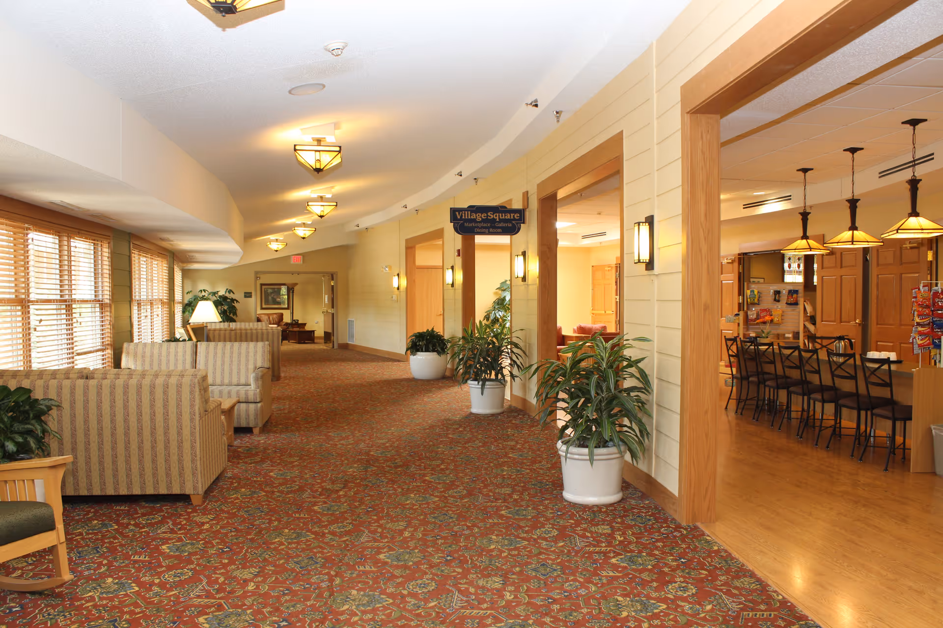 A well-lit hallway in a senior living facility with patterned carpet, several potted plants, and seating areas with striped armchairs and lamps along the left side. On the right side, there is an open entrance to a dining room area with a counter and bar stools, pendant lights, and snack displays. A sign above the entrance reads 'Village Square Marketplace - Galleria Dining Room.'
