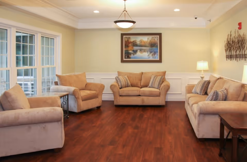 A cozy living room with three beige upholstered sofas arranged around the perimeter of the room. The room has wooden flooring, cream-colored walls with white wainscoting, and a large window on the left side letting in natural light. There is a framed landscape painting on the wall above the central sofa and two table lamps on either side providing warm lighting. A decorative metal wall art piece is mounted on the right wall.