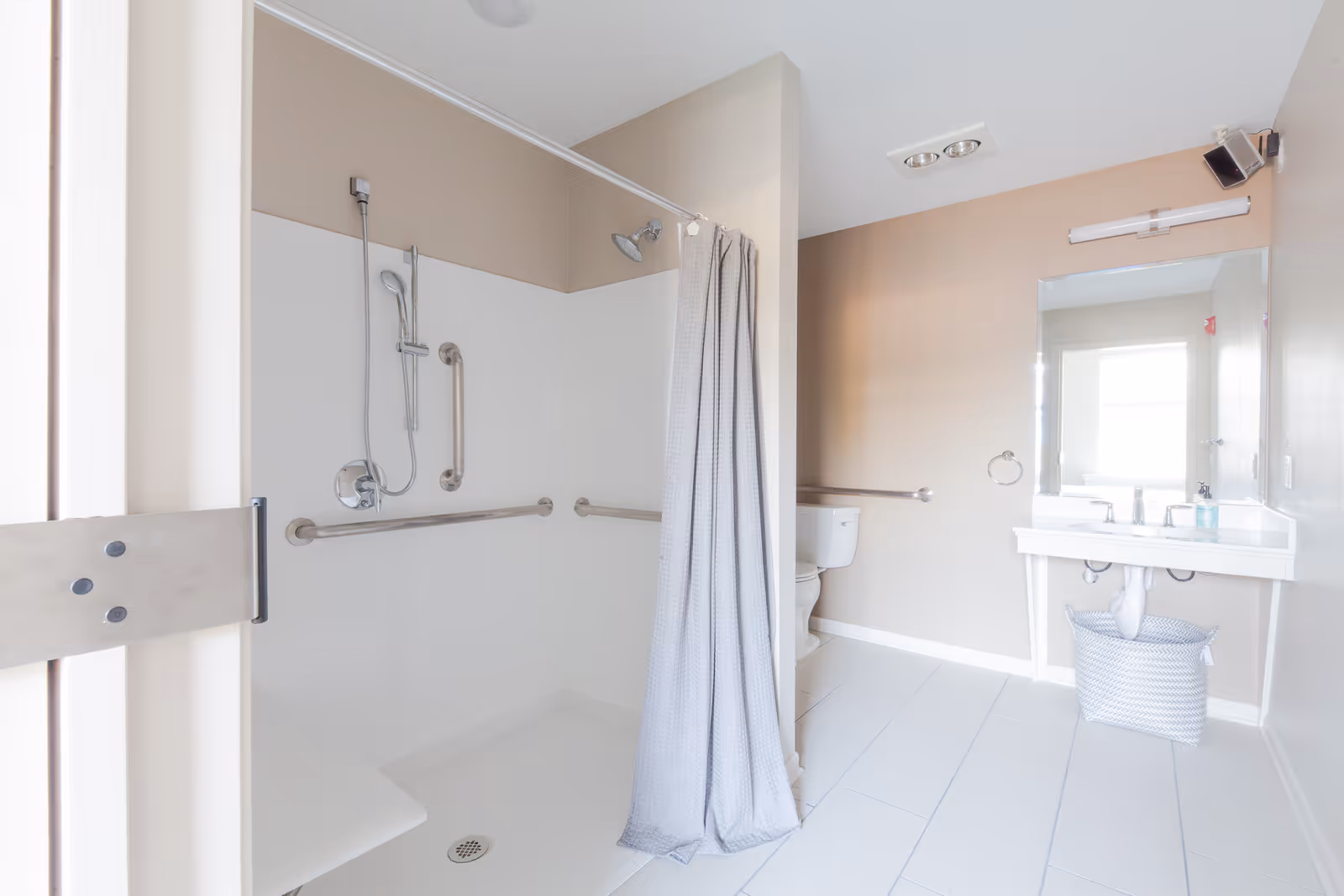 Bright and clean bathroom with a walk-in shower featuring grab bars and a handheld showerhead. The shower has a white curtain and a built-in bench. The bathroom also includes a white toilet, a white sink with a large mirror above it, a towel ring, and a woven basket underneath the sink. The walls are painted beige and the floor is tiled in white.