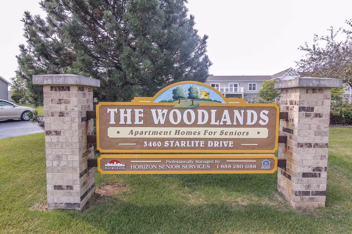 A large wooden sign for The Woodlands Senior Apartments, mounted between two brick pillars on a grassy area. The sign includes the address 3460 Starlite Drive and notes that the apartments are professionally managed by Horizon Senior Services with a contact number 1-888-280-1188. There are trees and residential buildings visible in the background.