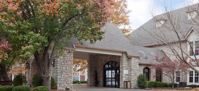 Stone and brick building entrance with an arched doorway, surrounded by trees with autumn foliage and shrubs, under a partly cloudy sky.