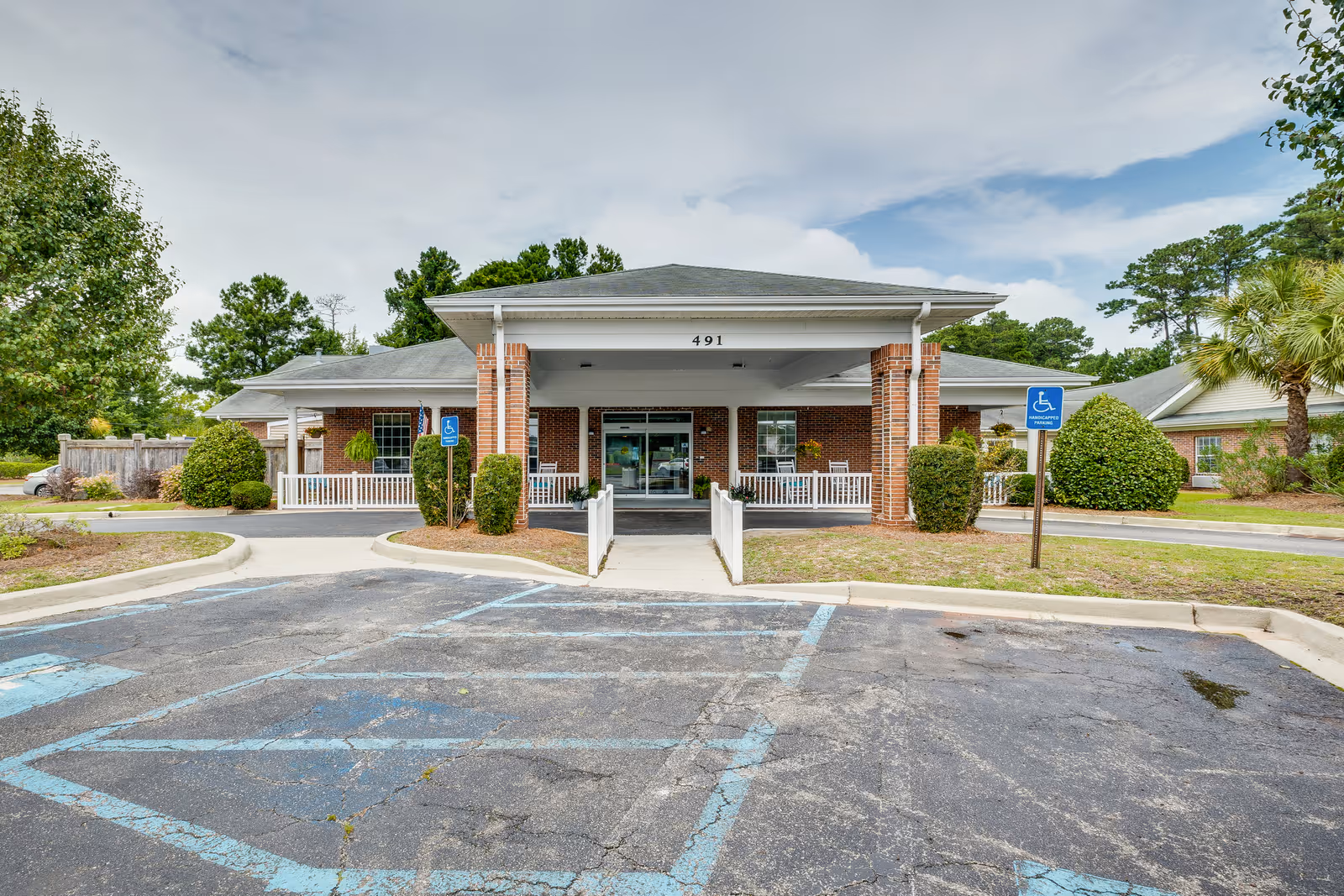 Front exterior view of a single-story brick building with a covered entrance, wheelchair accessible ramp, and designated handicapped parking spaces in front. The building is surrounded by greenery including bushes, trees, and a palm tree under a partly cloudy sky.