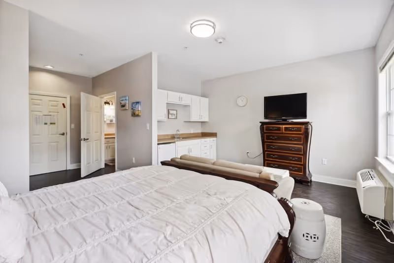 A bright and clean bedroom in a senior living facility featuring a large bed with white bedding, a small beige sofa at the foot of the bed, a wooden dresser with a flat-screen TV on top, a kitchenette with white cabinets and a sink, and a window letting in natural light. The room has light gray walls and dark wood flooring.