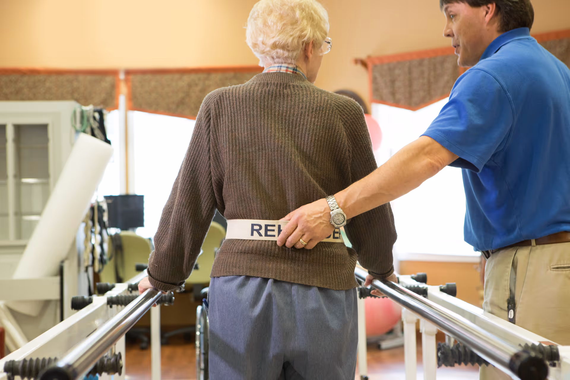 An elderly person using parallel bars for physical therapy with assistance from a caregiver in a rehabilitation setting.