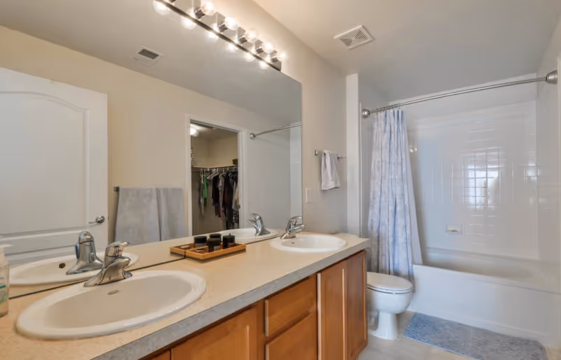 A bathroom with a double sink vanity featuring two faucets and a large mirror above. There is a bathtub with a shower curtain, a toilet, and a towel hanging on the wall. The bathroom has light-colored walls and a door leading to a walk-in closet with clothes hanging inside.
