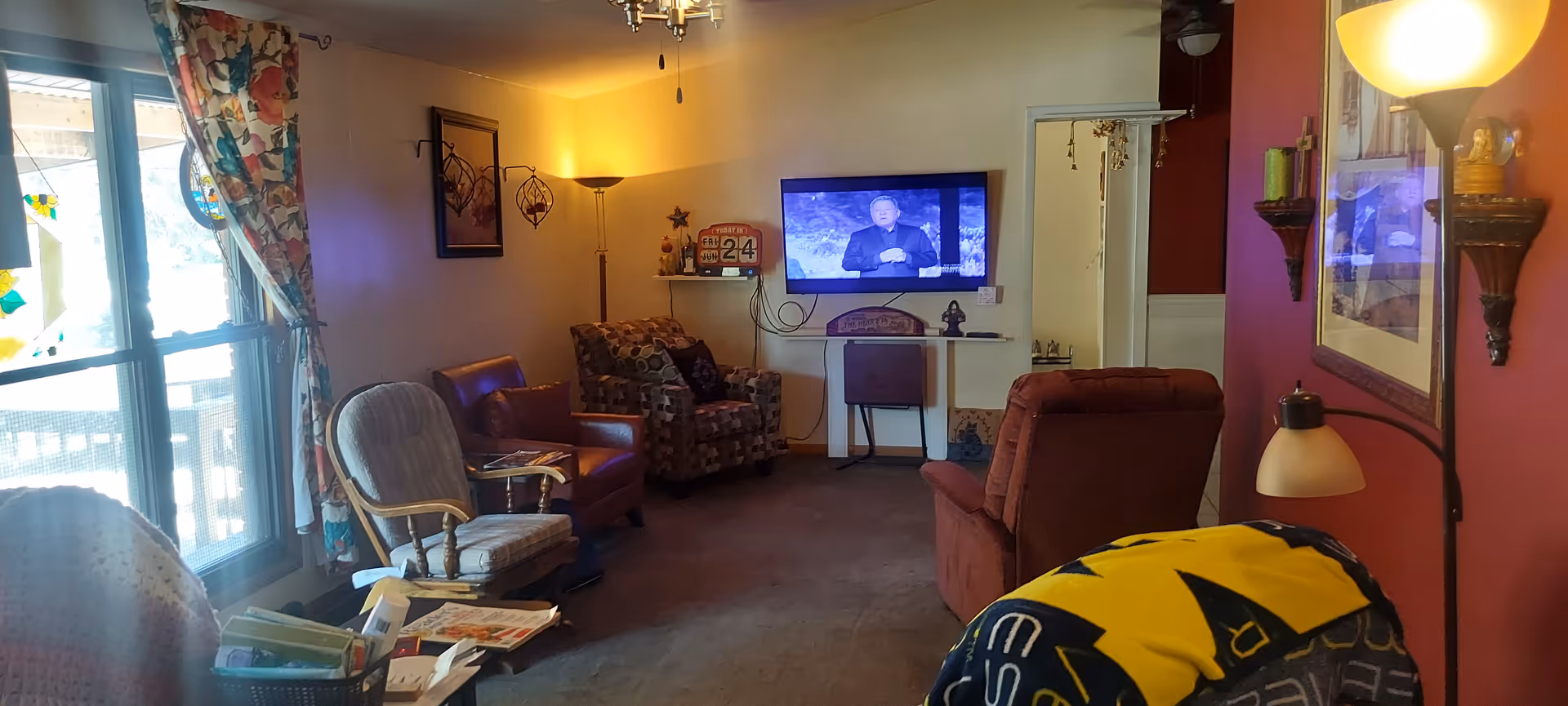 A cozy living room with several armchairs and a recliner arranged around a wall-mounted TV. The room has a large window with floral curtains letting in natural light. There are various decorative items including a floor lamp, wall art, and small shelves with ornaments. A blanket with a yellow and black pattern is draped over one chair.