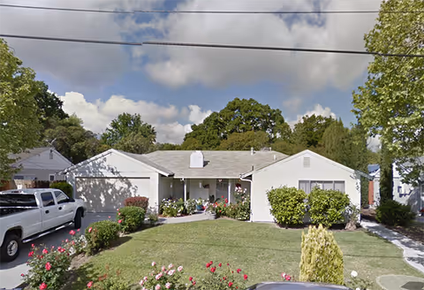 Single-story white residential building with a front lawn featuring various bushes and flowers. A white pickup truck is parked in the driveway on the left side. The sky is partly cloudy with trees in the background.