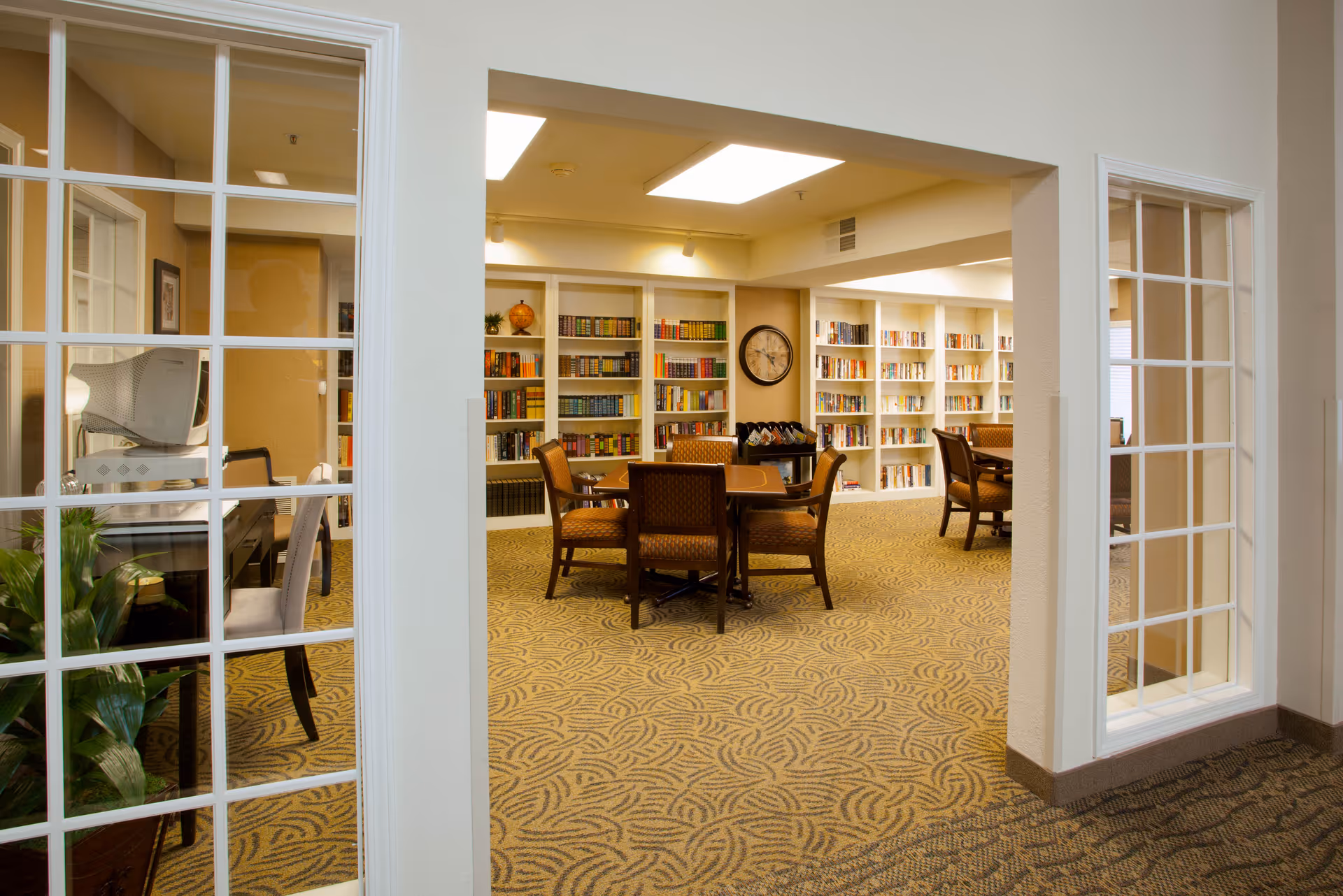 Interior view of a library or reading room in a senior living facility with bookshelves filled with books along the walls, several wooden tables with chairs arranged around them, and a clock mounted on the wall. The room is well-lit with ceiling lights and has patterned carpet flooring.