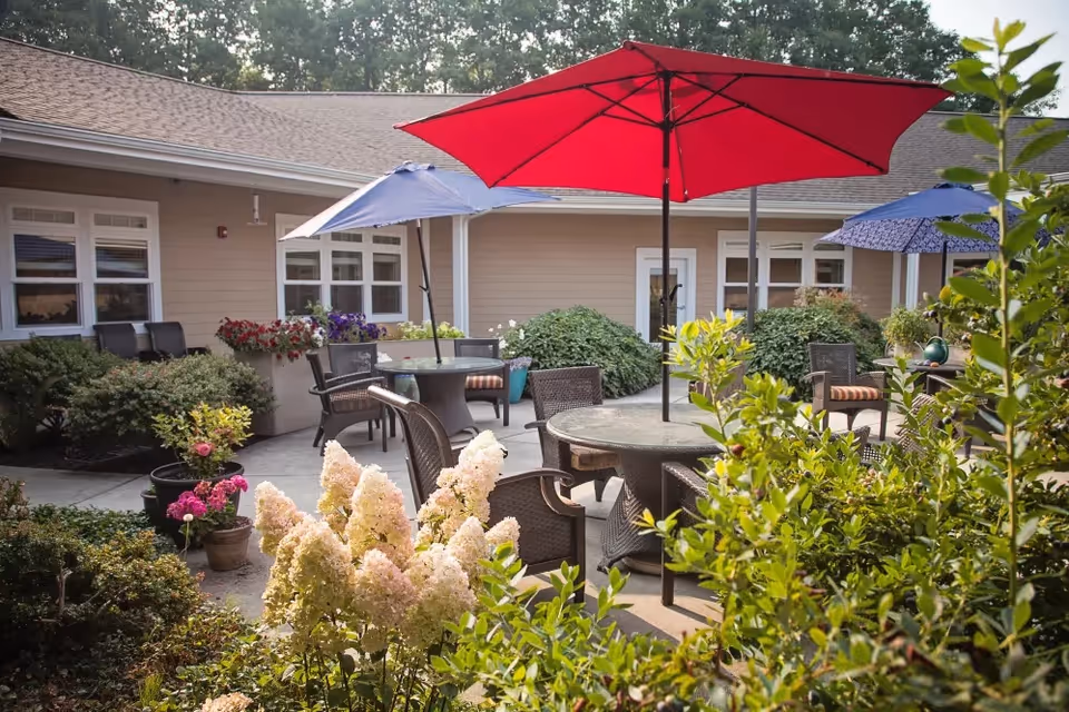 Sunny courtyard with patio tables and chairs, colorful umbrellas, and flowering shrubs outside a single-story memory care building.