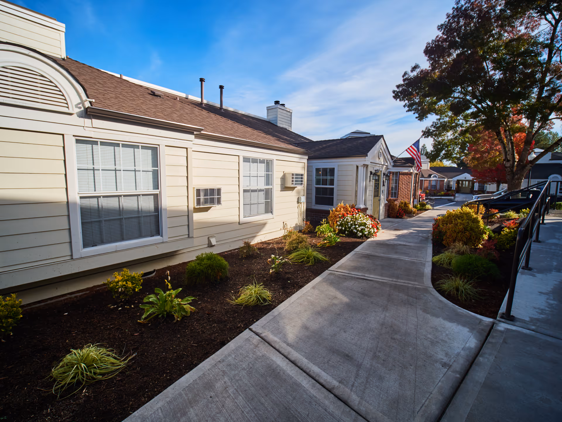Exterior view of a senior living facility building with beige siding and white-framed windows. A concrete walkway runs alongside the building, bordered by landscaped flower beds with various plants and shrubs. An American flag is mounted near the entrance, and trees with autumn foliage are visible in the background under a blue sky.