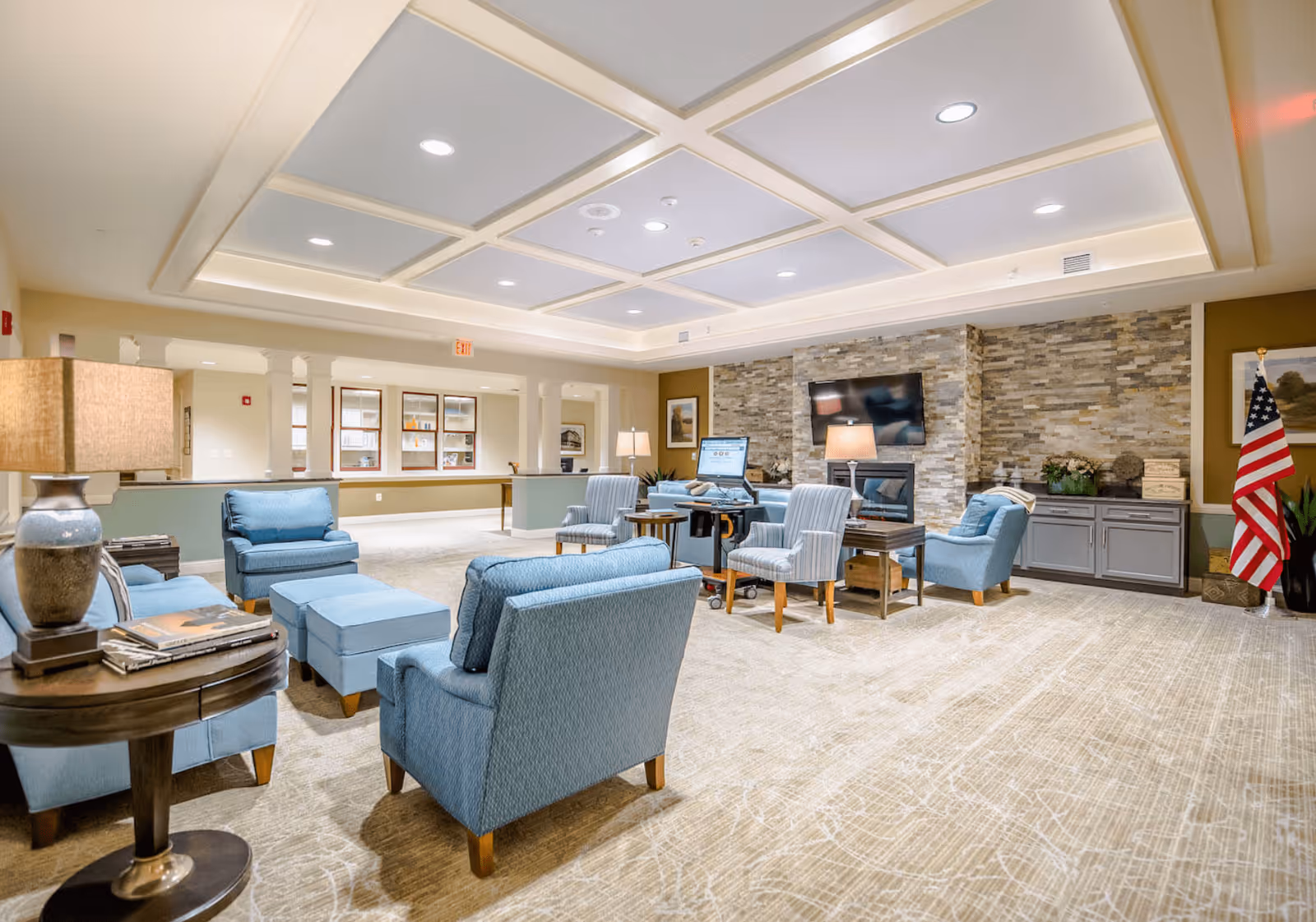 A spacious and well-lit living room area in a senior living facility featuring multiple blue upholstered armchairs and ottomans arranged around wooden side tables. The room has a coffered ceiling with recessed lighting, a stone accent wall with a mounted flat-screen TV above a fireplace, and an American flag in the corner. There are also cabinets and framed artwork on the walls, with a beige patterned carpet covering the floor.