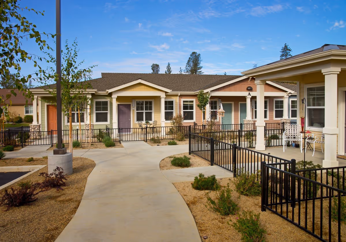 View of a single-story senior living community with multiple colorful units featuring front porches, small tables and chairs, surrounded by landscaped pathways and low black metal fencing under a clear blue sky.