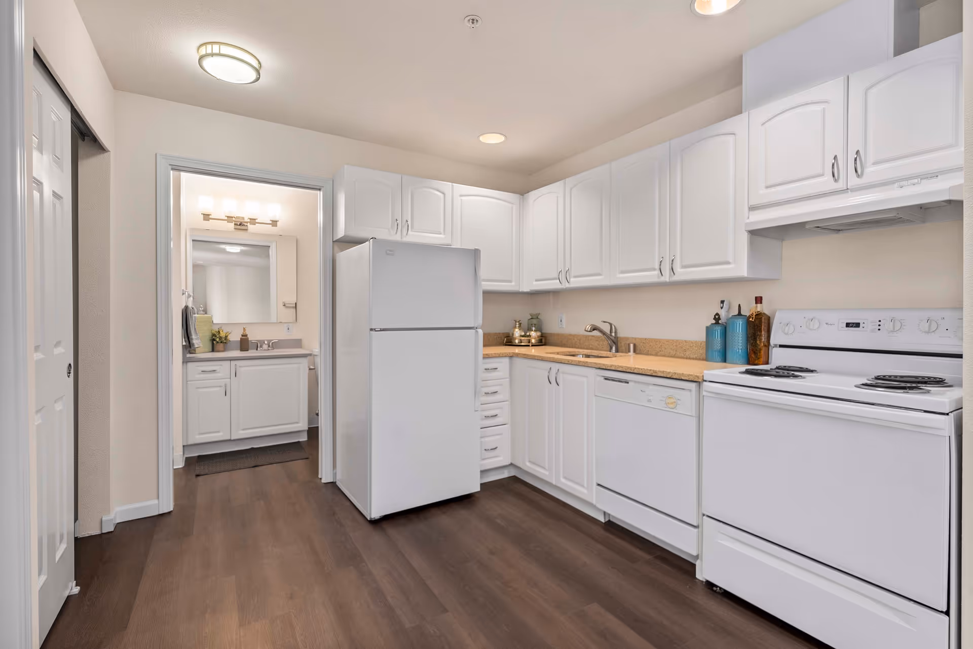 Well-lit kitchen with white cabinets, refrigerator, stove, dishwasher, sink, and a view into an adjacent bathroom vanity.