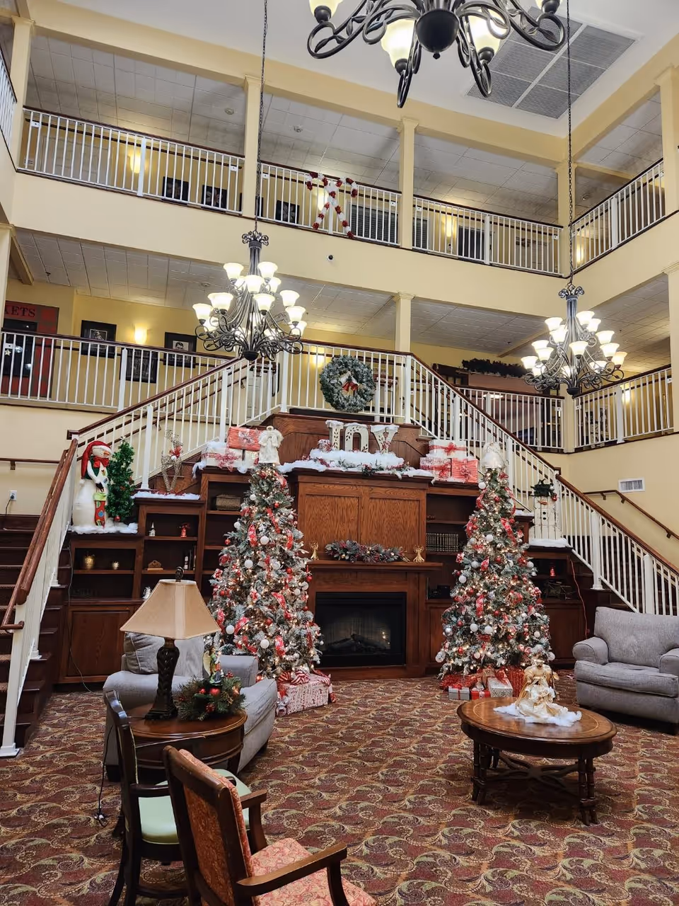 A spacious senior living community common area decorated for Christmas with two decorated Christmas trees, wrapped presents, a fireplace, a wreath, and festive ornaments. The room features a high ceiling with two large chandeliers, a patterned carpet, comfortable seating including armchairs and sofas, and a wooden staircase with white railings leading to an upper balcony.