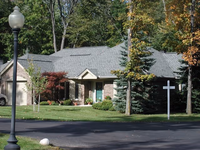 A single-story brick house with a gray shingled roof surrounded by trees and greenery. There is a green front door, a garage on the left side, and a white real estate signpost on the right side near the driveway. A streetlamp is visible in the foreground on the left side.