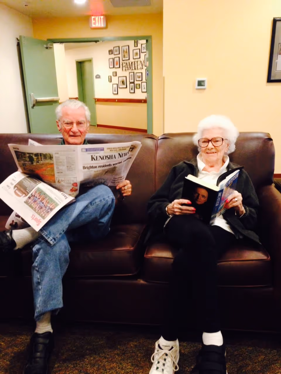 An elderly man and woman sitting on a brown leather couch in a living area. The man is reading a newspaper titled 'Kenosha News' and the woman is reading a book. The background shows a hallway with framed pictures on the wall and an exit sign above a door.