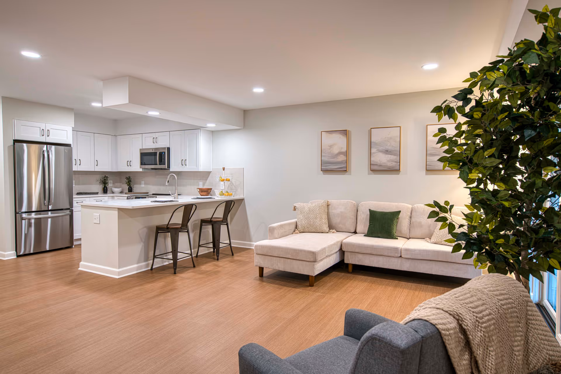 A modern open-concept living area featuring a kitchen with white cabinets, stainless steel refrigerator, microwave, and a kitchen island with two metal bar stools. Adjacent to the kitchen is a beige sectional sofa with decorative pillows, three framed artworks on the wall, a gray armchair with a knitted throw, and a large green plant near a window.