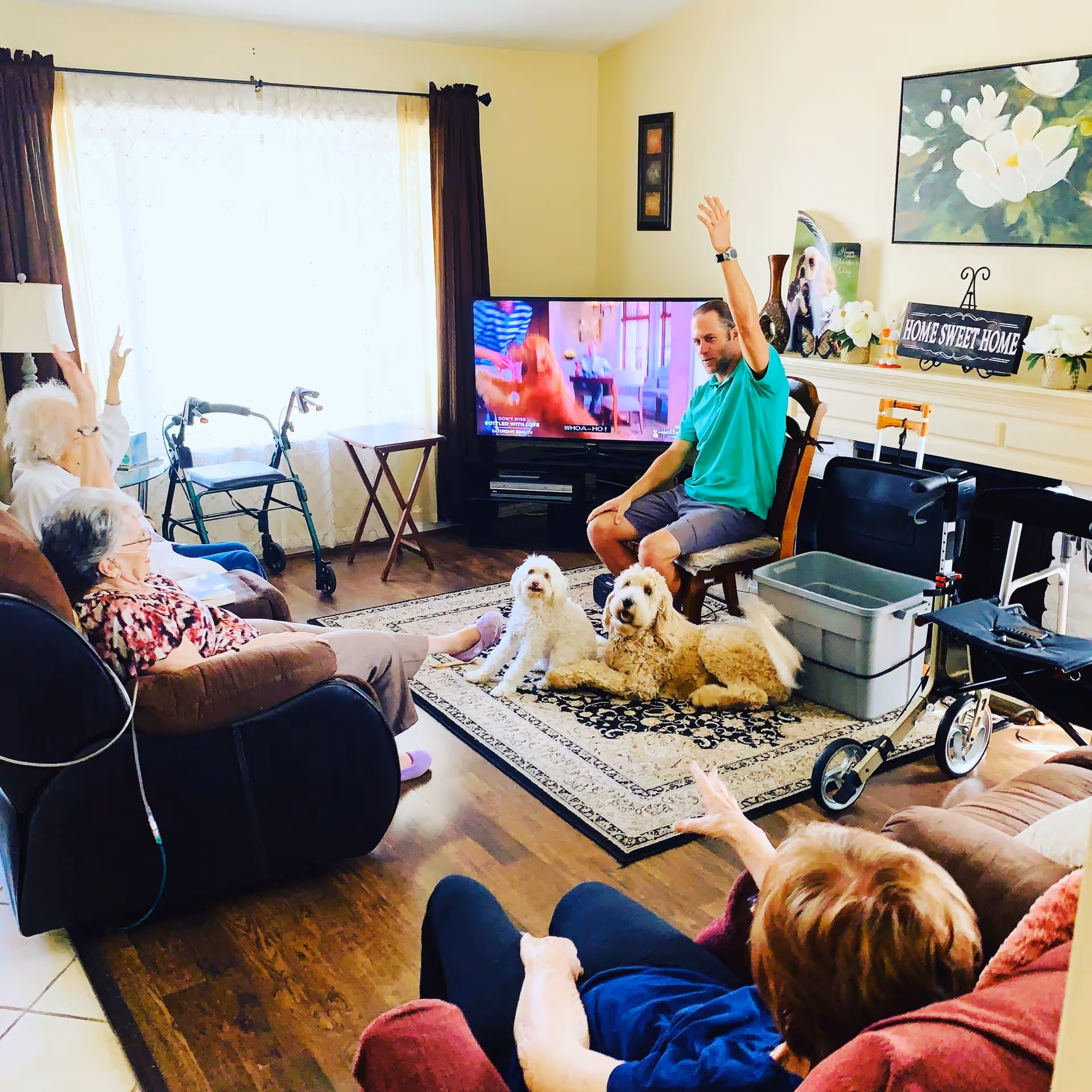 A cozy living room scene in a senior care facility with three elderly women seated on couches and chairs, a man sitting on a chair with his hand raised, and two dogs lying on a patterned rug. The room has a TV, a walker, a wheelchair, and home decor including a 'Home Sweet Home' sign and floral artwork on the wall.