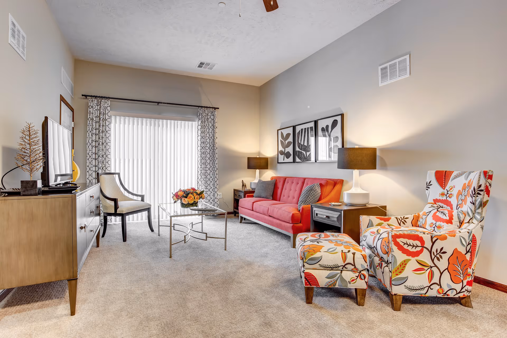 A bright and cozy living room with a red sofa, a floral patterned armchair with matching ottoman, a glass coffee table with a flower arrangement, a wooden TV stand with a flat-screen TV, and a white chair near a large window with vertical blinds and patterned curtains. Two table lamps with black shades illuminate the room, and three framed botanical prints hang on the wall above the sofa.