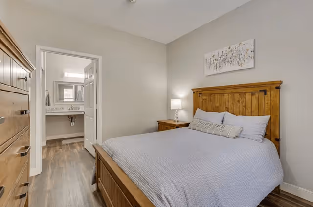 A bedroom with a wooden bed frame and matching nightstand and dresser. The bed is made with light gray bedding and decorative pillows. A lamp is on the nightstand, and a piece of artwork hangs above the bed. The room has light-colored walls and wood flooring. An open door reveals a bathroom with a sink and mirror.