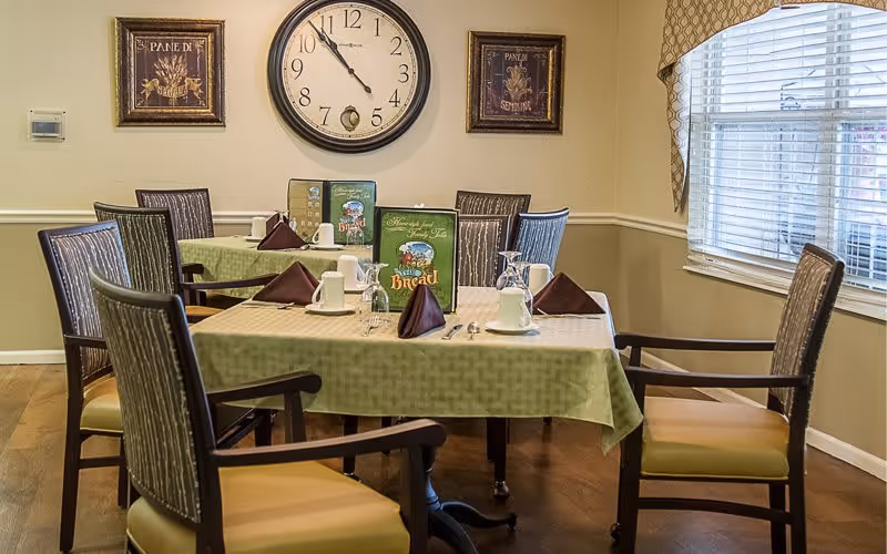 A dining room with a table set for six people, featuring green tablecloths, folded brown napkins, white cups, glasses, and menus. The room has wooden chairs with cushioned seats, a large clock on the wall, two framed pictures, and a window with blinds and patterned curtains.