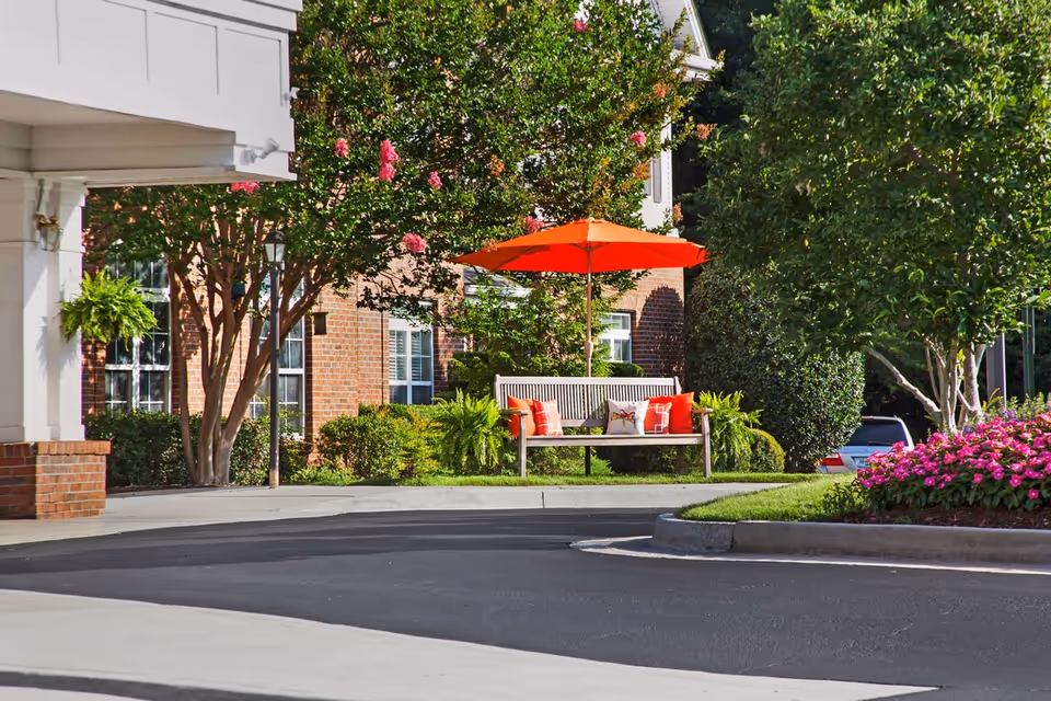 Outdoor seating area at Brighton Gardens of Dunwoody featuring a wooden bench with colorful cushions under an orange umbrella, surrounded by green shrubs, flowering plants, and trees near a brick building.