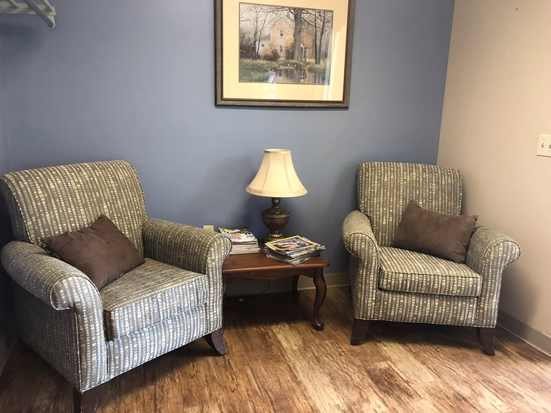Two patterned armchairs with brown cushions are positioned on either side of a wooden side table with a lamp and stacks of magazines. The wall behind is painted blue and beige, with a framed picture of a house and trees hanging above the table. The floor is wooden.