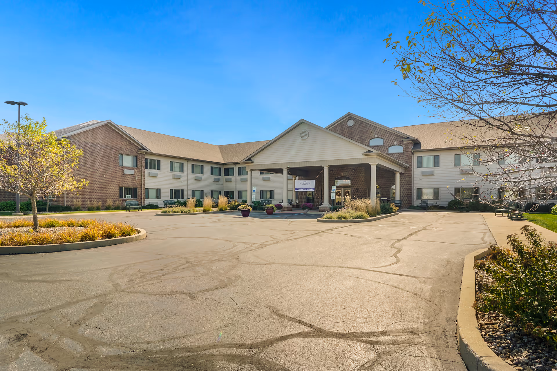 Exterior view of a two-story senior living facility building with a covered entrance, surrounded by a paved driveway and landscaped areas with trees and shrubs under a clear blue sky.