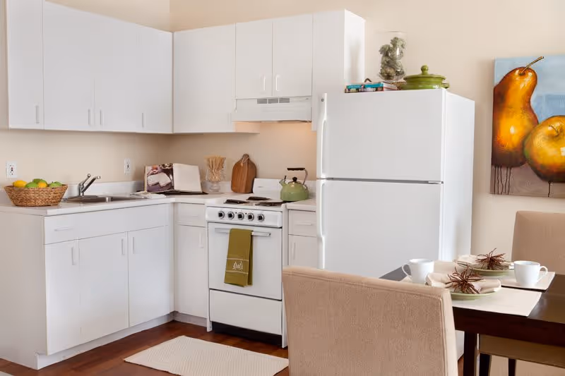 A small kitchen area with white cabinets, a white refrigerator, a white stove with a green kettle on it, and a sink. There is a basket of fruit on the counter, a green towel hanging on the oven door, and a dining table with beige chairs set with white cups and plates. A painting of pears hangs on the wall.