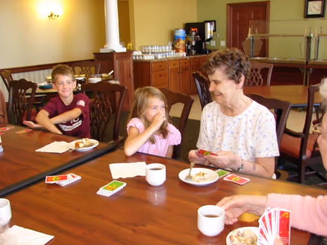 An elderly woman and two children sit at a dining table in a community dining room, playing cards and drinking coffee.