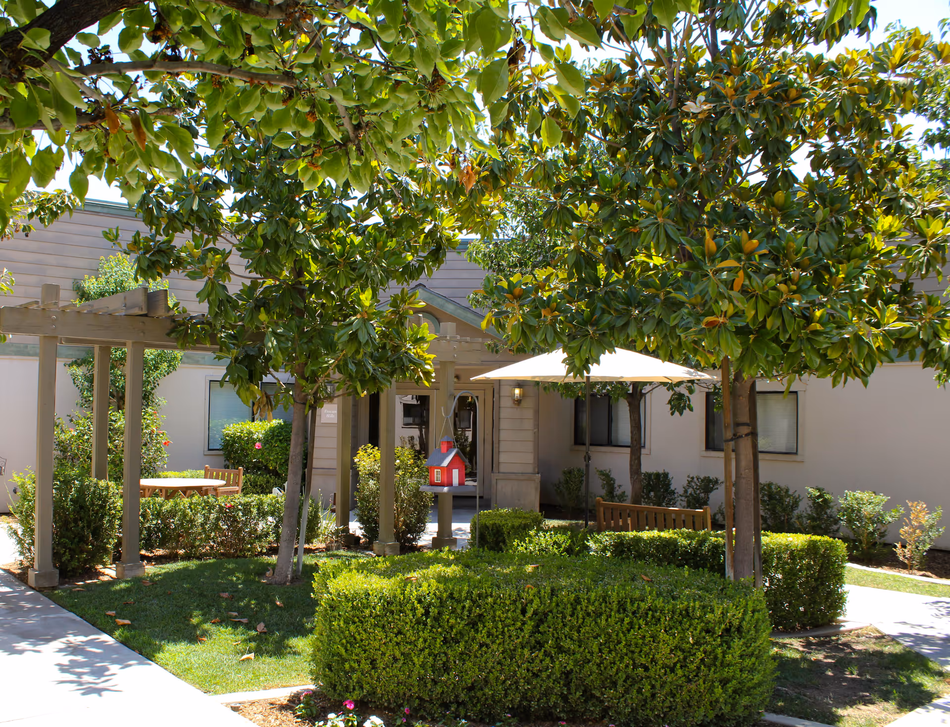 Outdoor garden area at a senior living facility with green trees, trimmed hedges, a wooden pergola, benches, a table with an umbrella, and a small decorative red birdhouse on a post near the entrance of a beige building.