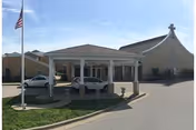 Entrance area of a facility with a covered drop-off zone supported by white columns. Two cars are parked under the covered area. An American flag is flying on a flagpole to the left. The building has a large, curved roof section on the right side.