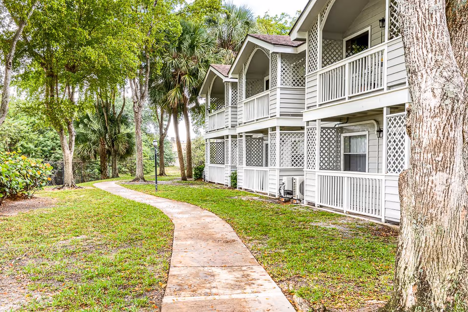 Outdoor view of a senior living facility with a paved walkway curving through a grassy area lined with trees. On the right side, there is a two-story building with white railings and latticework on the balconies and porches.