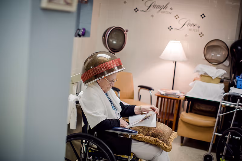 An elderly woman sitting in a wheelchair under a hair dryer in a salon area, reading a magazine. The room has chairs, a lamp, and wall decor with the words 'Laugh Often' and 'Love Much'.