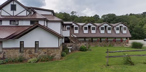 Exterior front view of a white two-story building with brown metal roofs, dormer windows, stone foundation, and a grassy lawn with a wooden fence.