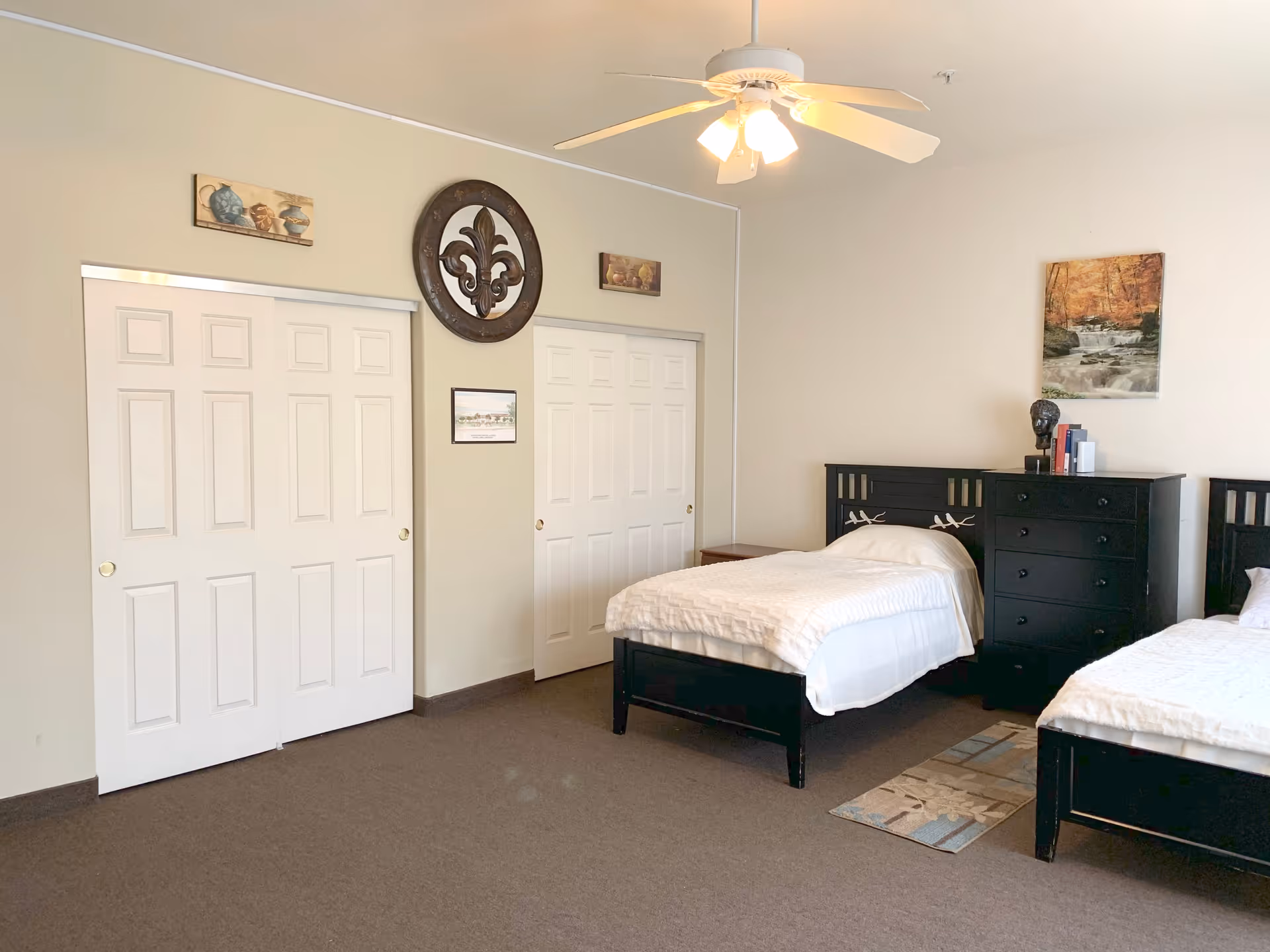 A bedroom with two single beds featuring black wooden headboards and white bedding. Between the beds is a black dresser with books and a decorative item on top. The room has beige walls with two white sliding closet doors, a ceiling fan with lights, and wall decorations including a circular ornamental piece and framed pictures.