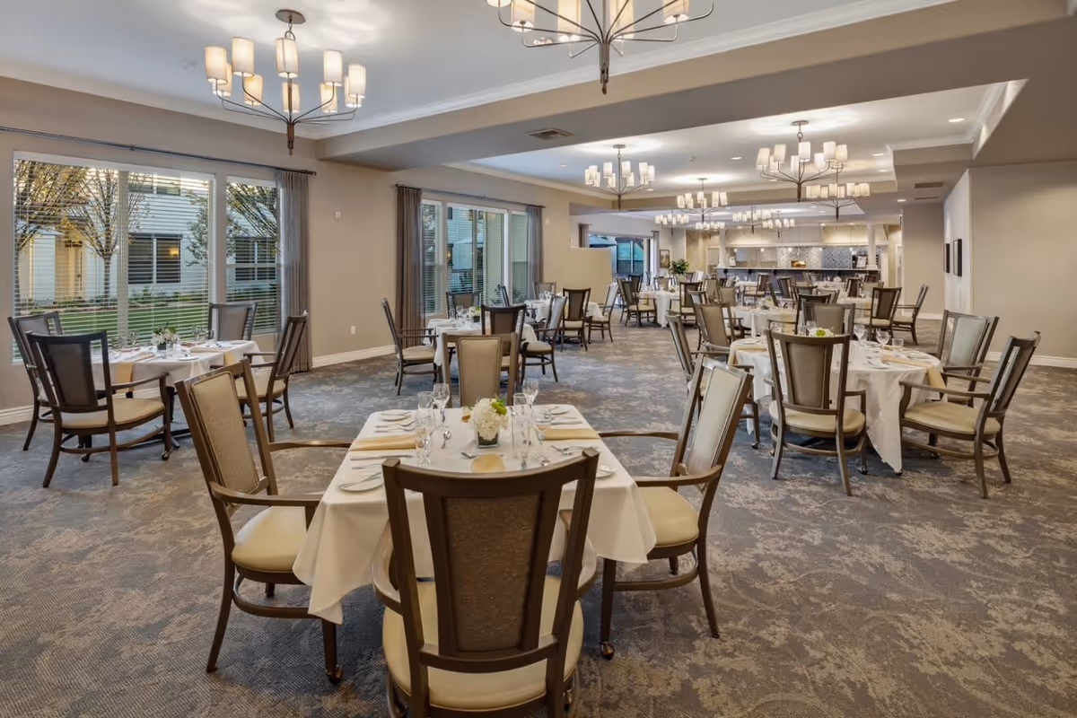 A spacious dining room with multiple round tables covered with white tablecloths, each set with plates, glasses, and silverware. The room features large windows with curtains, modern chandeliers hanging from the ceiling, and a carpeted floor with a subtle pattern. The dining area extends towards a kitchen or serving area in the background.