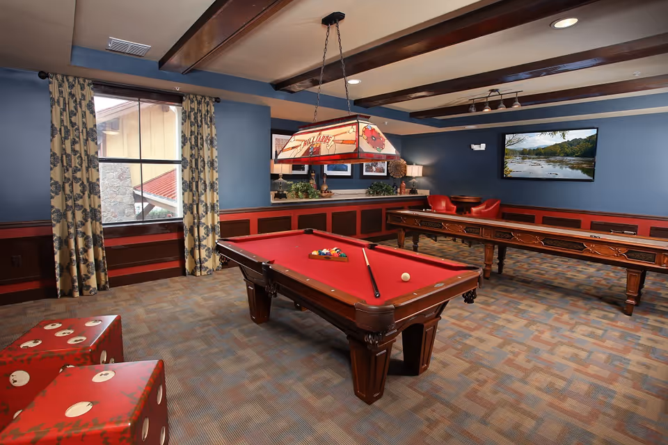 Game room interior featuring a red-felt pool table, shuffleboard, seating, patterned curtains and a wall-mounted TV.