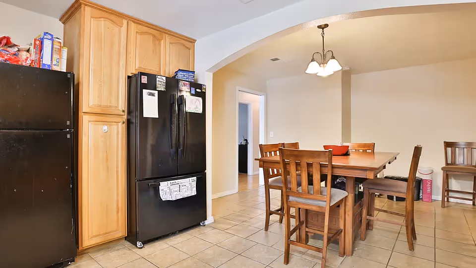 A kitchen and dining area with two black refrigerators, wooden cabinets, and a wooden dining table surrounded by six chairs. The floor is tiled, and a light fixture hangs above the dining table. There is an archway leading to another room.