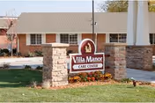 Outdoor view of the entrance sign for Villa Manor Care Center, featuring a stone base with a maroon and white sign displaying the facility's name, surrounded by grass and some landscaping with flowers.
